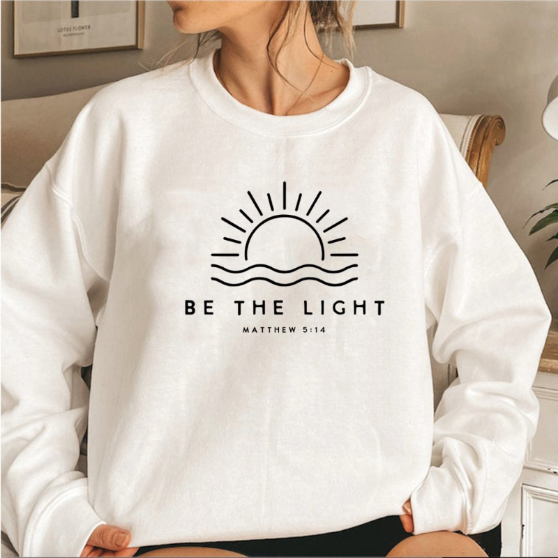 Femme portant un sweat blanc avec un motif de soleil et l'inscription "Be the Light Matthew 5:14", mode décontractée, intérieur moderne.