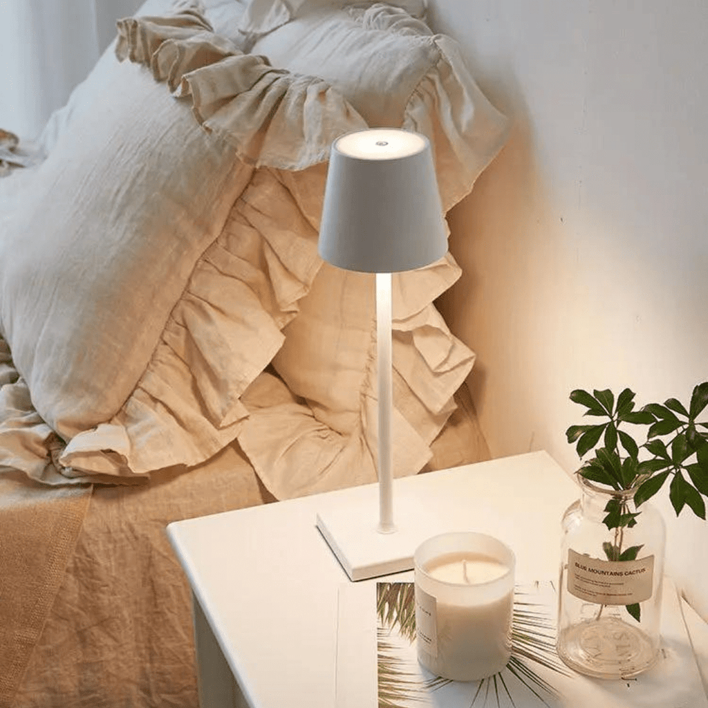 Lampe de chevet moderne blanche sur table de nuit, à côté d'un lit avec coussins beiges et une bougie parfumée, ambiance cosy et élégante.