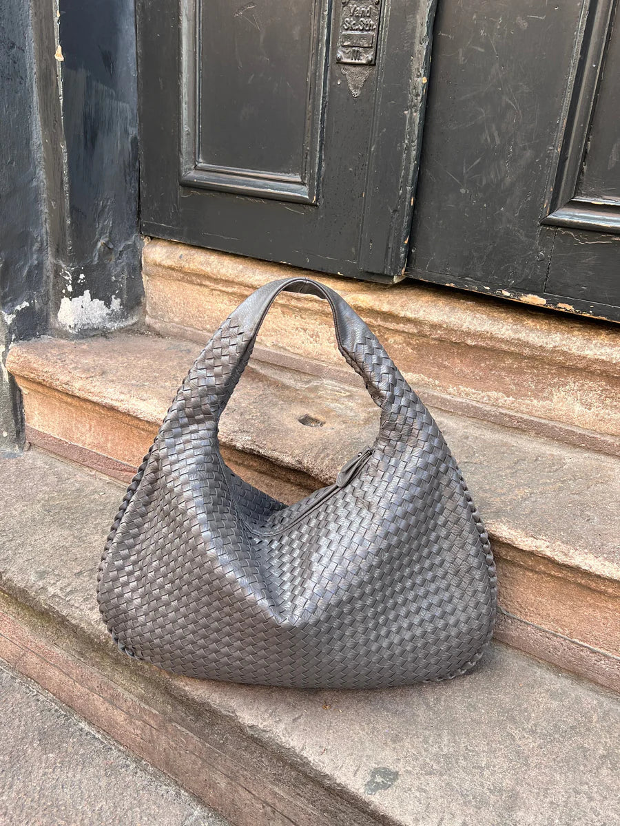 Sac à main en cuir tressé gris posé sur des marches en pierre devant une porte noire. Accessoire mode élégant et tendance.