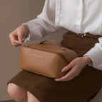 Femme ouvrant une élégante trousse de toilette en cuir marron, vêtue d'une chemise blanche et jupe marron. Accessoire mode chic et pratique.