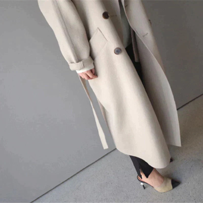 Manteau long beige élégant, ceinturé, avec boutons, porté par une personne en pantalon noir et talons. Mode automne-hiver, style chic et moderne.