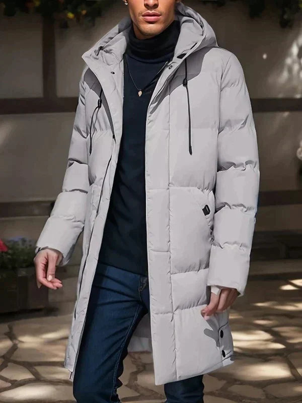 Homme portant un manteau d'hiver gris clair avec capuche, style moderne, sur fond urbain. Mode masculine, vêtements chauds, tendance hiver.