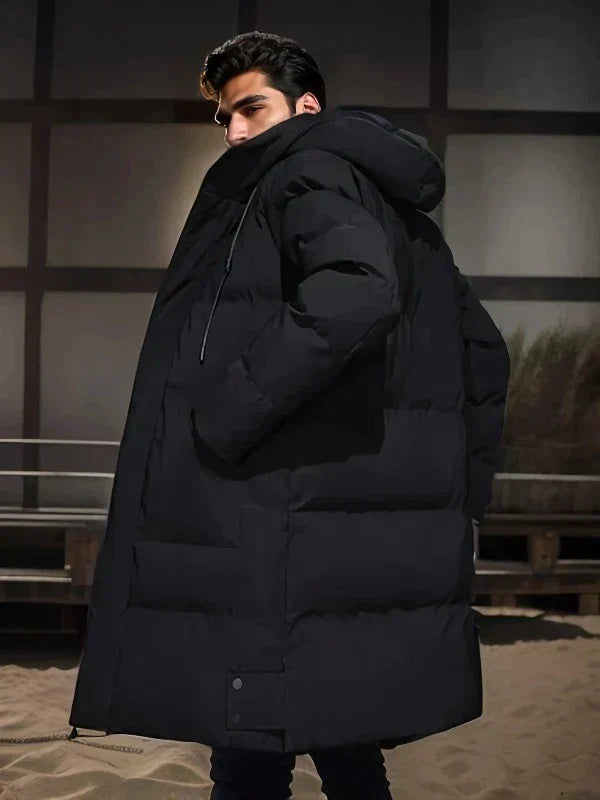Homme portant un manteau doudoune noir épais avec capuche, style hiver, mode masculine, arrière-plan urbain.