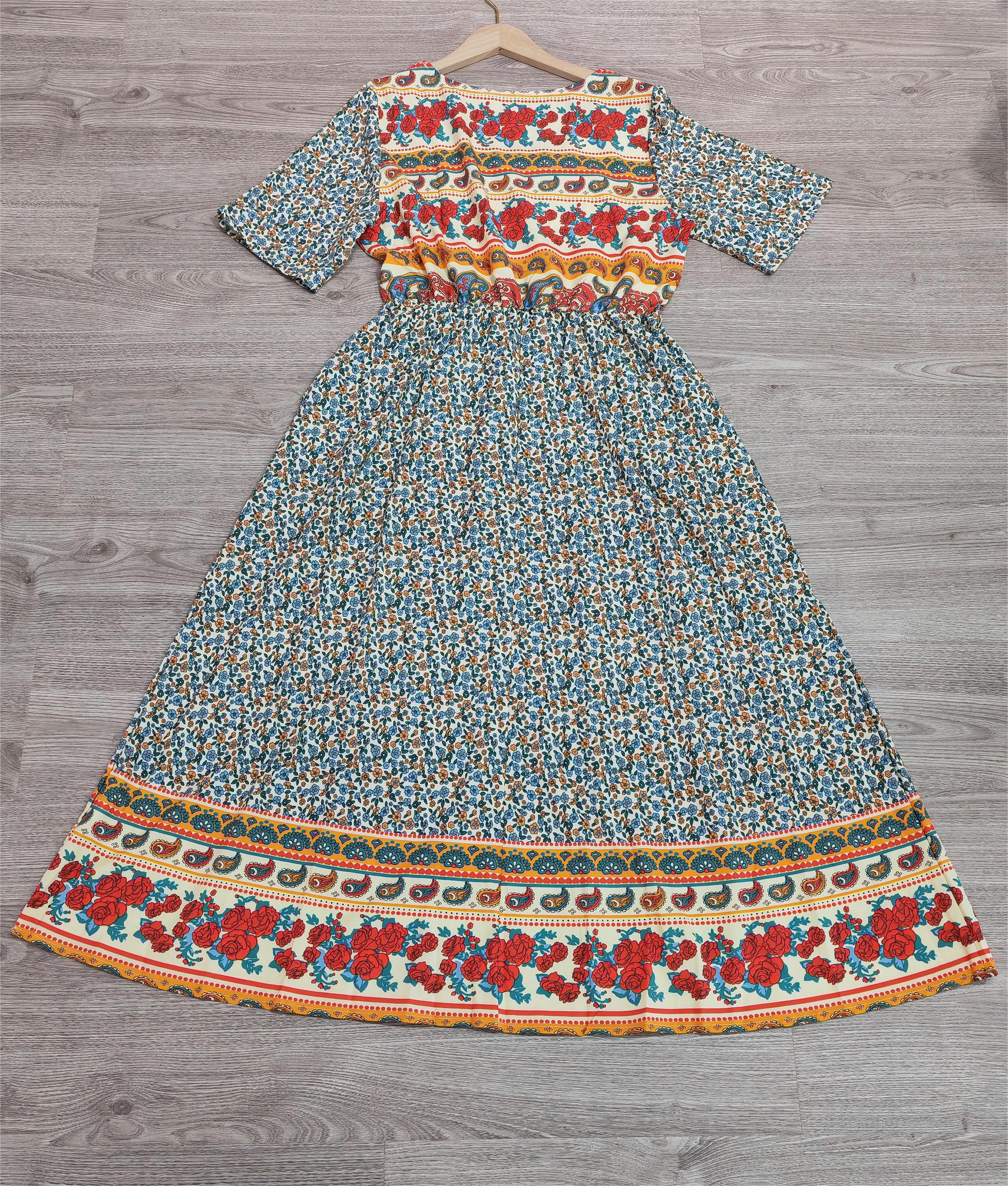 Robe longue bohème à motifs floraux colorés, manches courtes, fond bleu, accrochée sur un cintre en bois, posée sur un sol en bois gris.