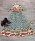 Robe longue bohème à motifs floraux colorés sur fond gris, manches courtes, accompagnée de bottines noires et épis de blé décoratifs.