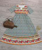 Robe longue bohème à motifs floraux colorés sur fond bleu, posée sur parquet gris, avec panier en osier et épis de blé. Mode féminine estivale.
