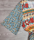 Robe bohème à motifs floraux colorés, manches évasées, fond en bois. Mode féminine, style vintage, tendance été.