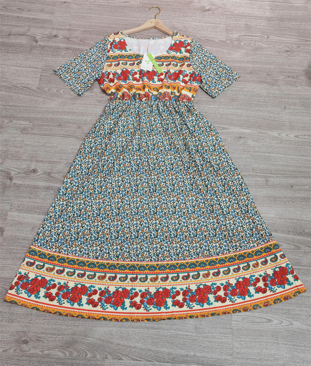 Robe longue bohème à motifs floraux multicolores, manches courtes, fond bleu, accrochée sur un cintre en bois, posée sur un sol en bois gris.
