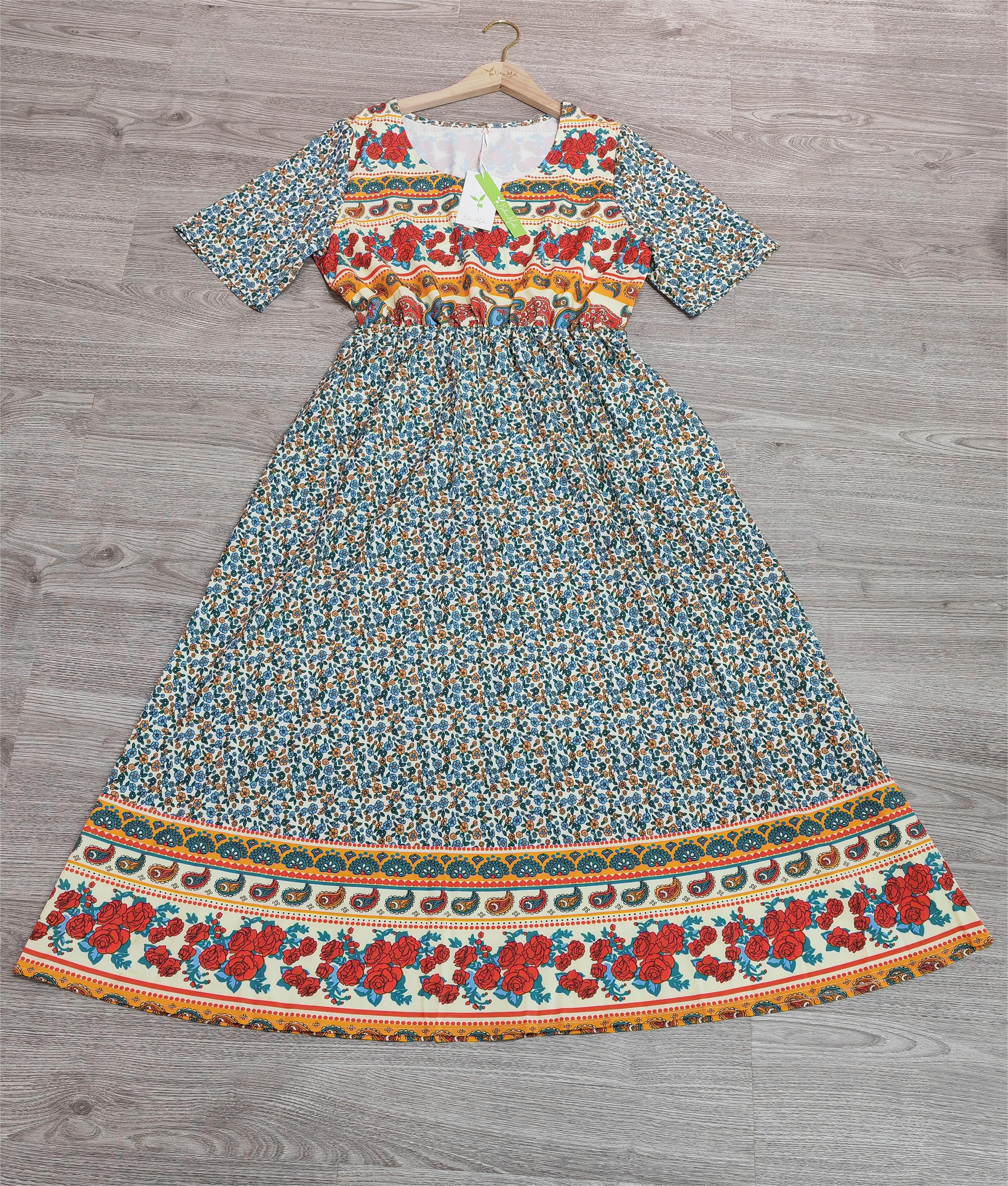 Robe longue bohème à motifs floraux multicolores, manches courtes, fond bleu, accrochée sur un cintre en bois, posée sur un sol en bois gris.