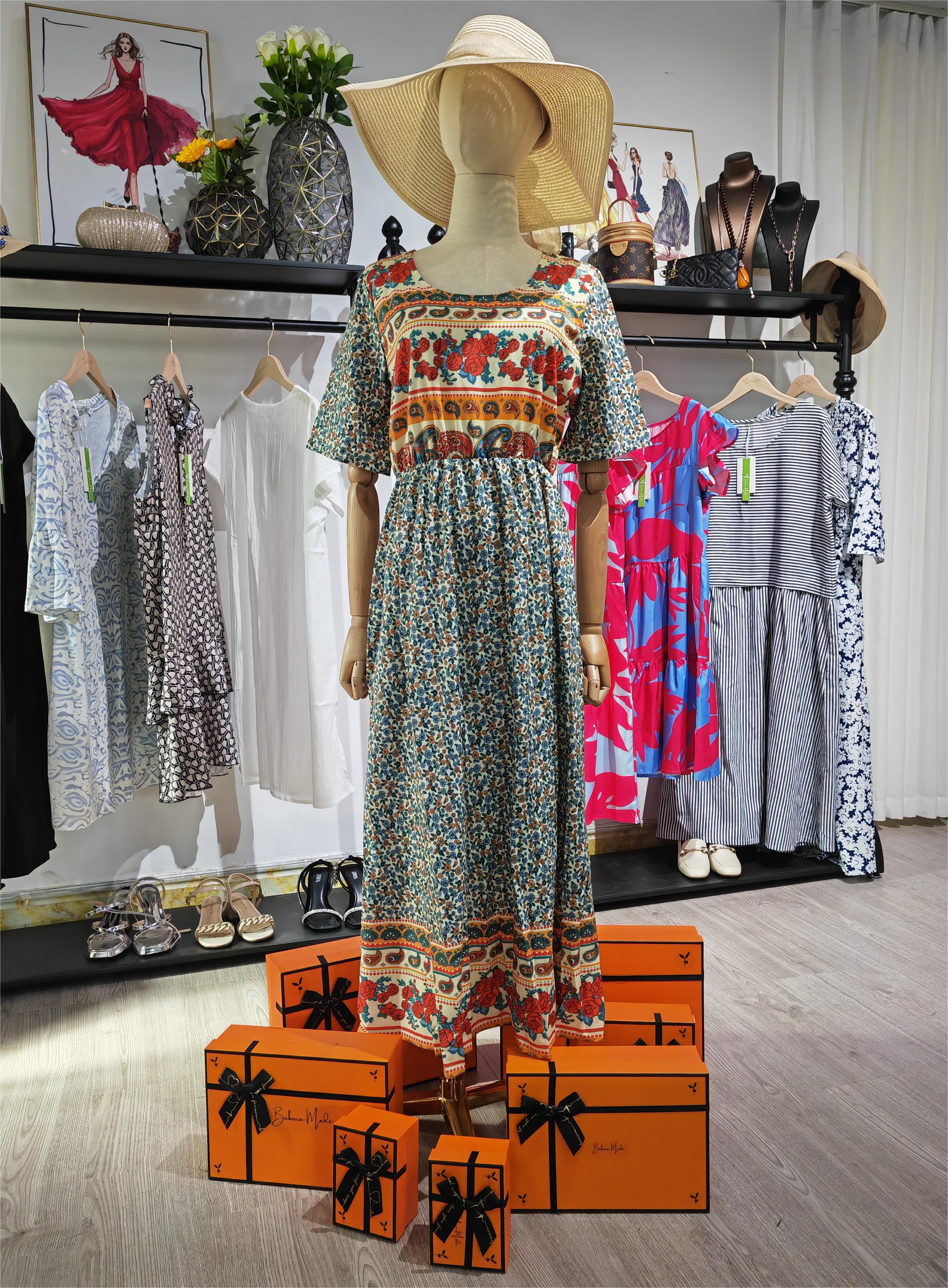 Mannequin portant une robe bohème colorée avec chapeau de paille, entouré de boîtes cadeaux orange et vêtements en arrière-plan dans une boutique élégante.