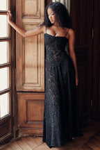Femme en robe longue noire en dentelle, posant près d'une fenêtre en bois, lumière naturelle, élégance, mode féminine, style chic.
