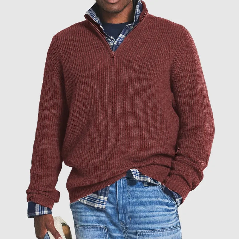 Homme portant un pull rouge bordeaux à col zippé, superposé sur une chemise à carreaux bleue, avec un jean décontracté. Mode automne-hiver tendance.
