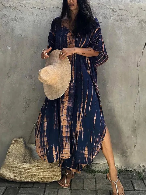 Femme en robe longue tie-dye bleue et beige, tenant un chapeau de paille, sandales marron, sac en osier au sol, mur en arrière-plan.