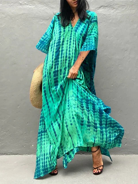 Robe longue bohème verte et bleue, motif tie-dye, portée par une femme avec des sandales noires, tenant un chapeau de paille. Mode estivale décontractée.