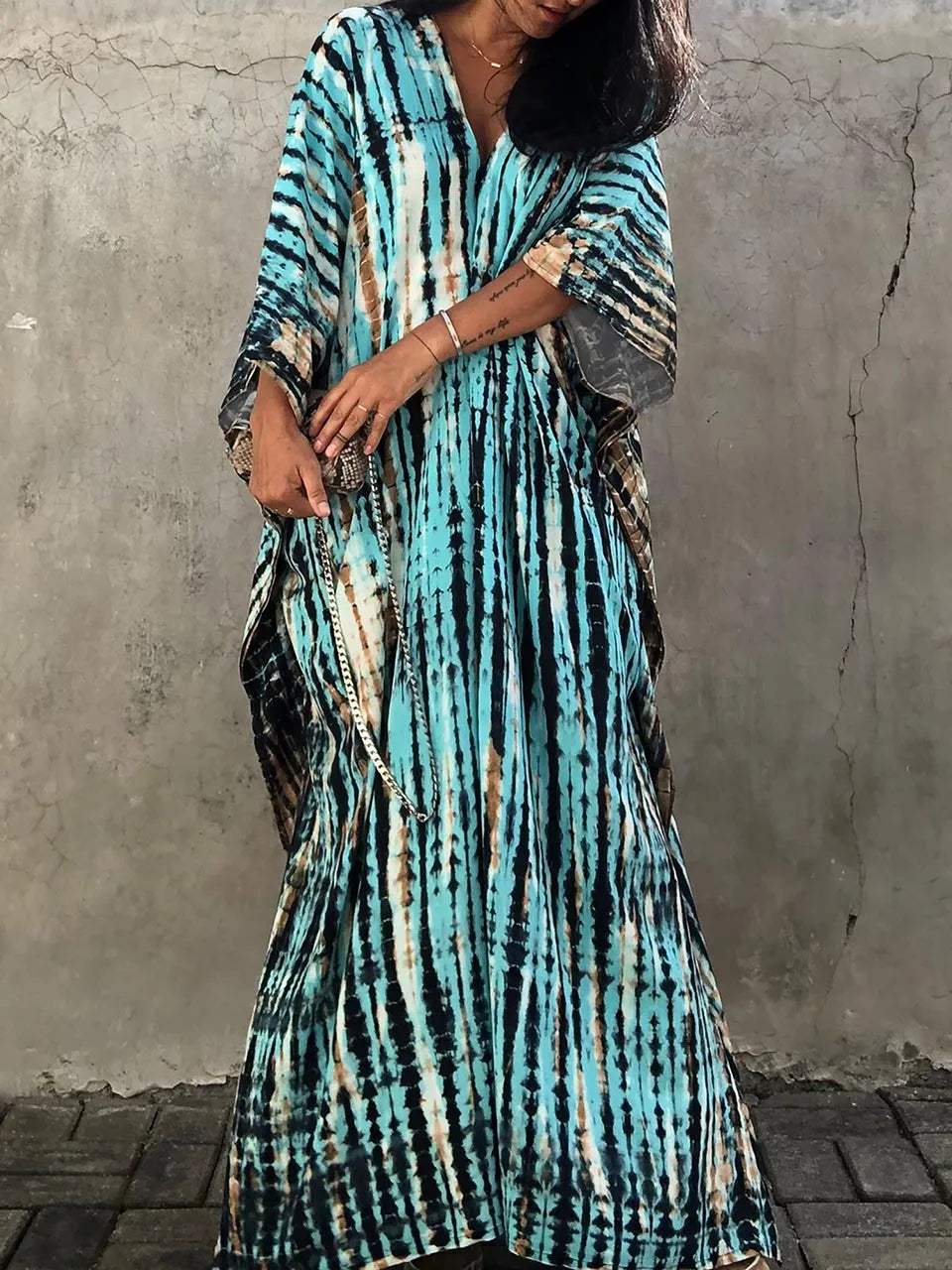Robe longue à motifs tie-dye bleu et marron, manches larges, portée par une femme devant un mur gris. Mode bohème chic, tendance été.