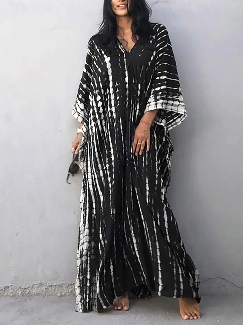 Robe longue noire et blanche à motifs tie-dye, manches larges, portée par une femme debout contre un mur gris, style bohème chic.