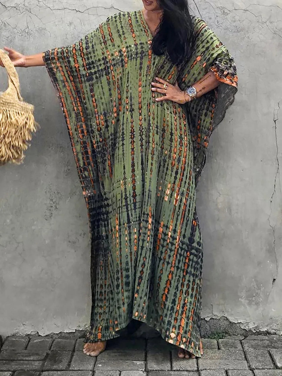 Robe longue bohème verte à motifs ethniques, manches larges, portée par une femme tenant un sac en paille. Mode estivale, style décontracté.