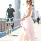 Femme en robe longue rose et blanche, souriante, marchant sur un pont ensoleillé, cycliste flou en arrière-plan, style estival élégant.