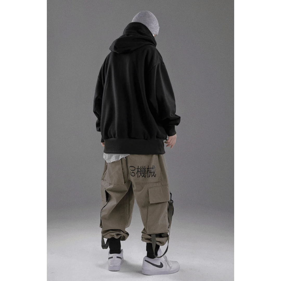 Homme en sweat à capuche noir, pantalon cargo beige avec motifs japonais, baskets blanches. Mode urbaine, style streetwear moderne.
