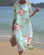 Robe longue fleurie femme, manches trois-quarts, fond bleu clair, motif floral rose, plage en arrière-plan, style estival élégant.