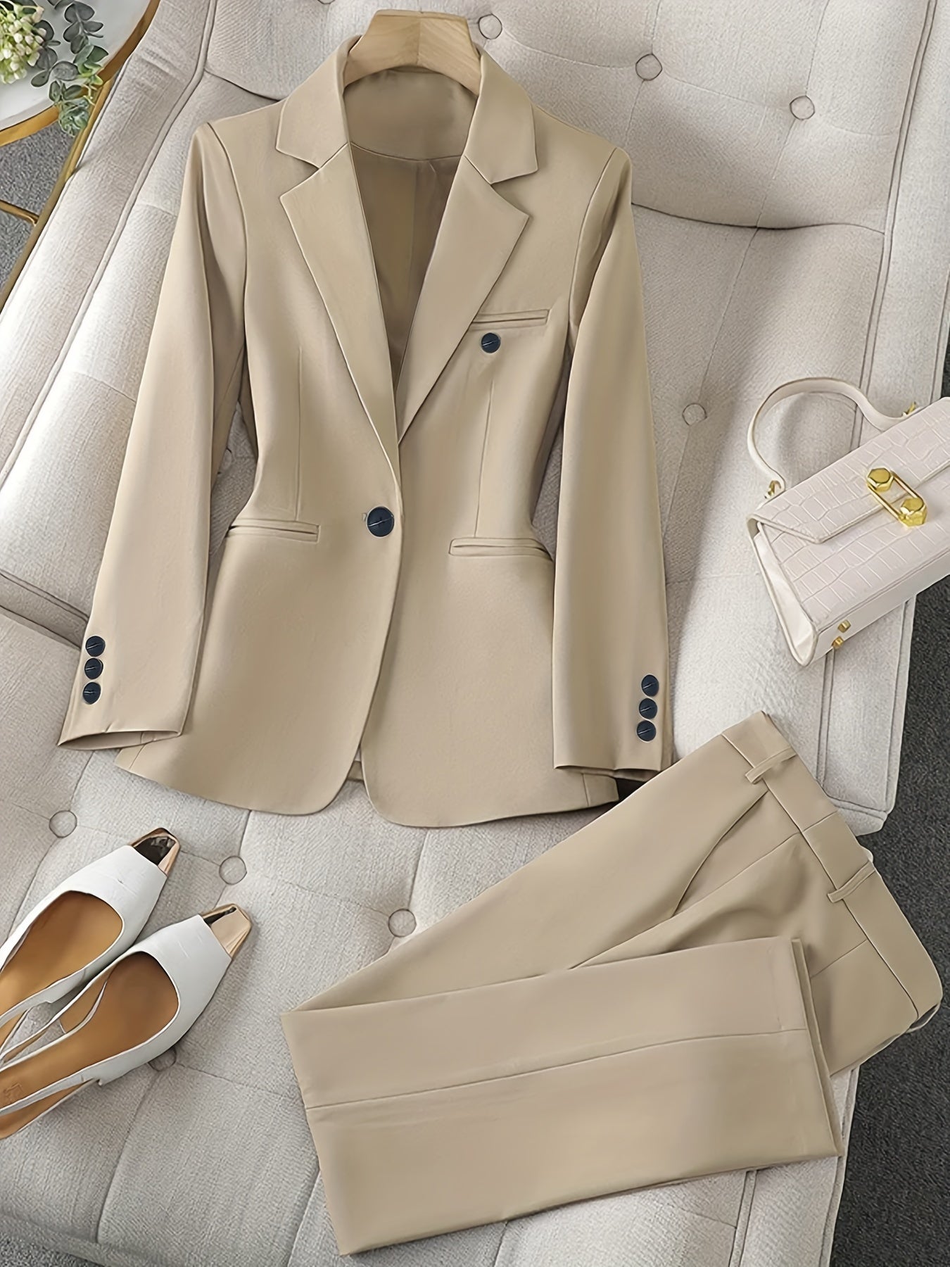 Costume beige élégant pour femme, veste et pantalon assortis, posés sur un canapé beige. Sac à main blanc et chaussures blanches à talons dorés.