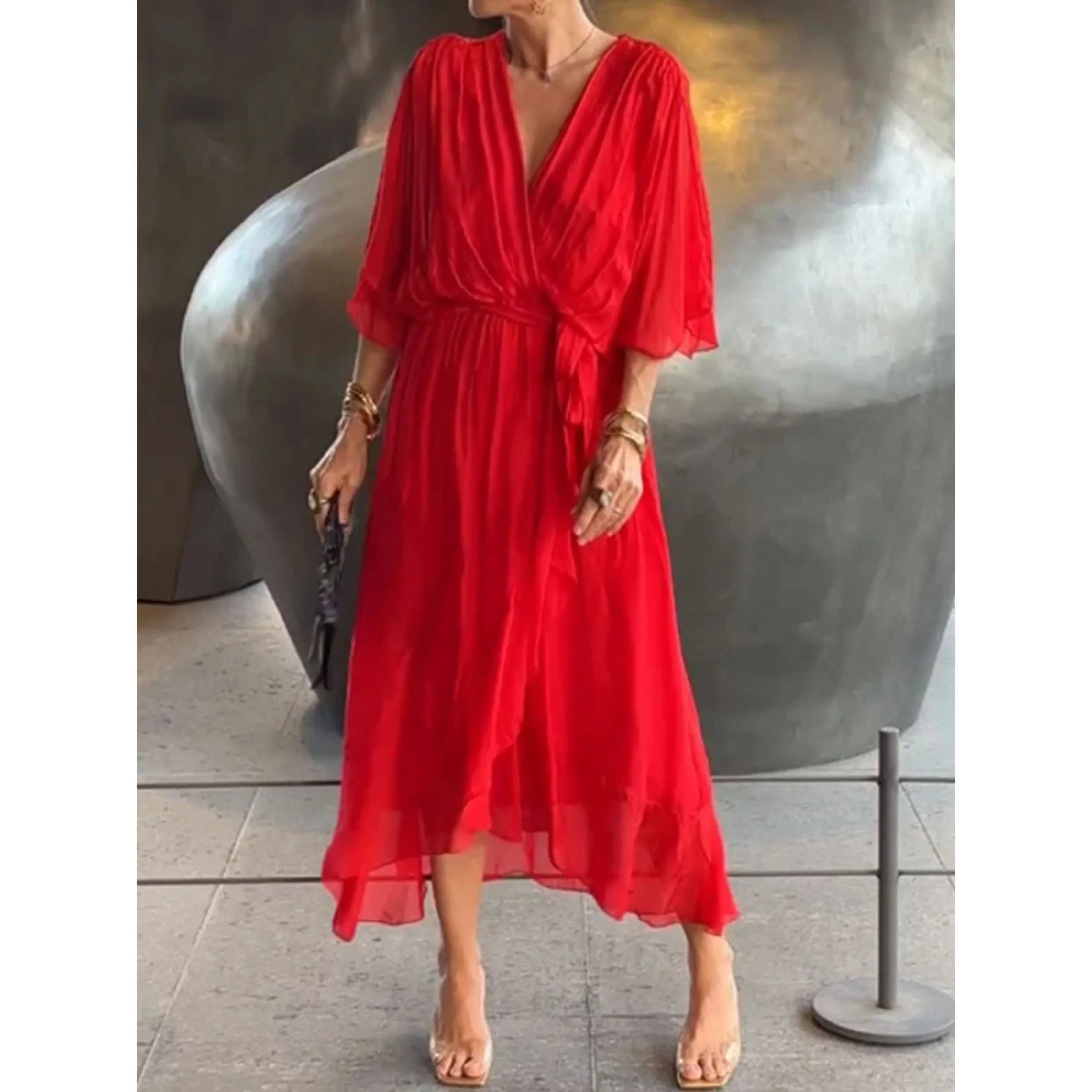 Robe rouge élégante en mousseline, manches trois-quarts, ceinture nouée, portée par une femme avec accessoires dorés, devant une sculpture moderne.