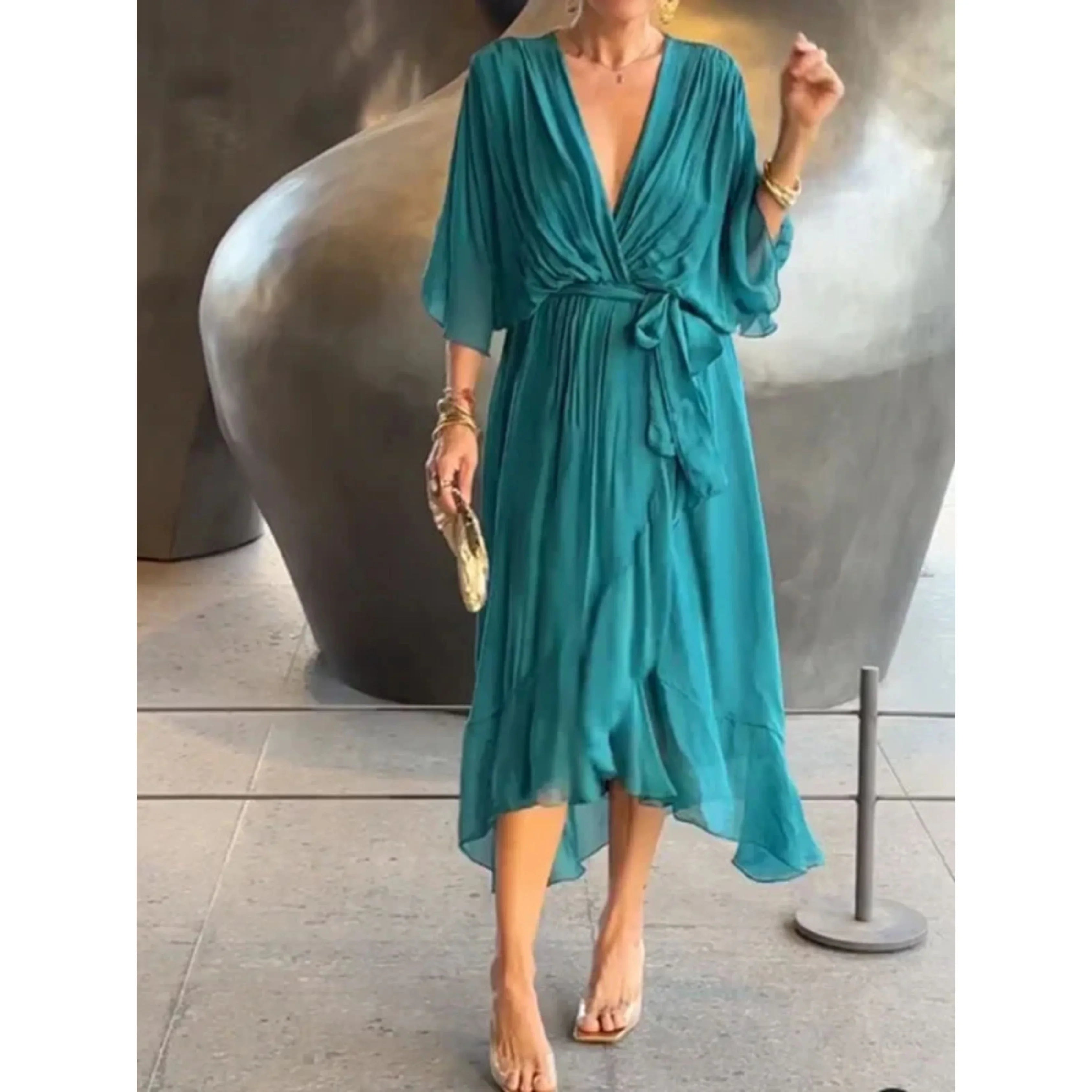 Robe longue bleu turquoise élégante avec décolleté plongeant, manches amples et ceinture nouée, portée par une femme avec accessoires dorés.
