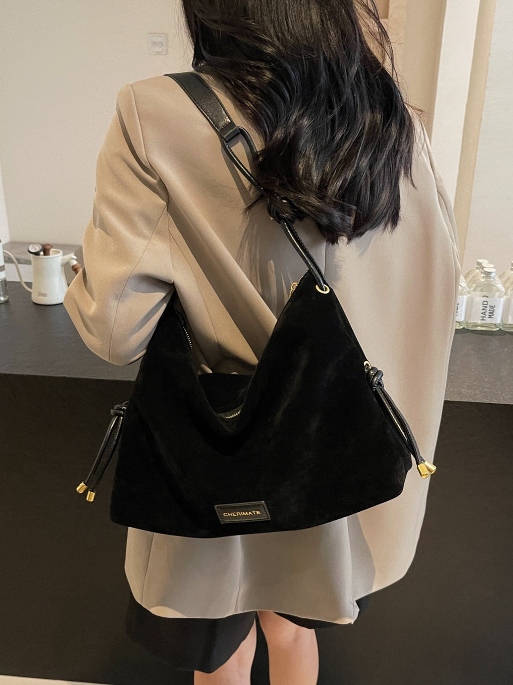 Femme portant un sac à main noir en daim, élégant et moderne, avec une étiquette dorée, sur un manteau beige. Accessoire de mode chic et tendance.