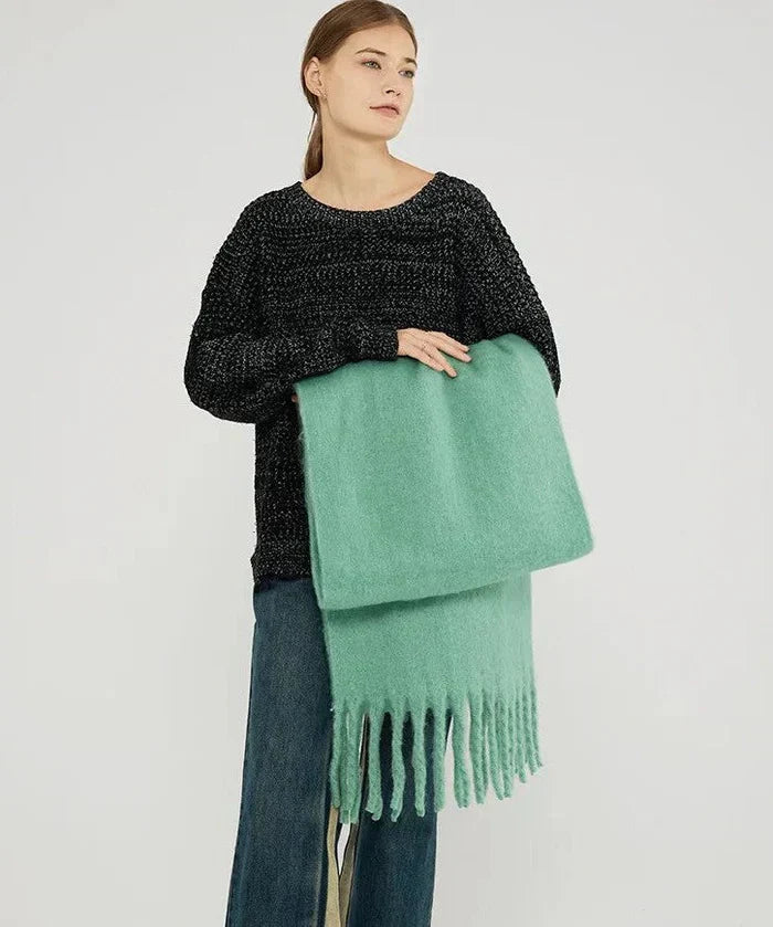 Femme portant un pull noir et tenant une écharpe verte en laine avec franges, mode automne-hiver, style décontracté, fond blanc.