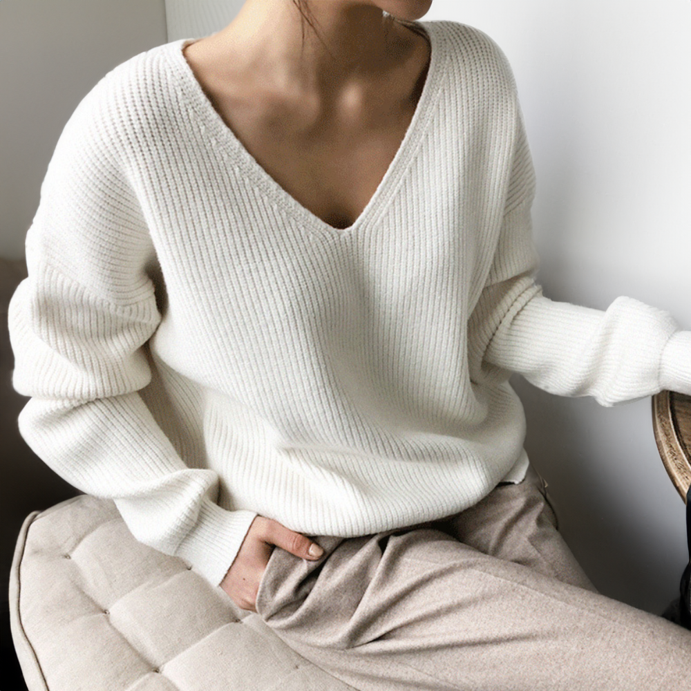 Femme portant un pull en maille blanc à col en V, assise sur un canapé beige. Mode automne-hiver, style décontracté et élégant.