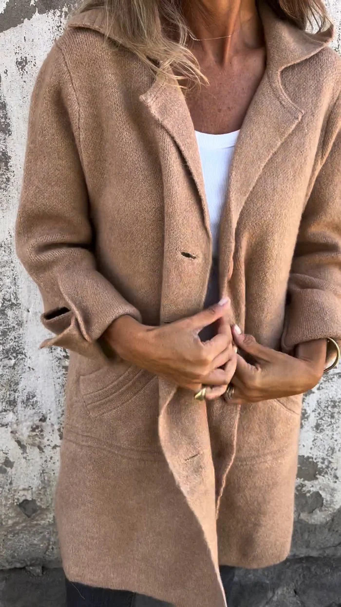 Femme portant un manteau en laine beige, manches retroussées, devant un mur texturé. Mode automne, style décontracté, tendance vestimentaire.