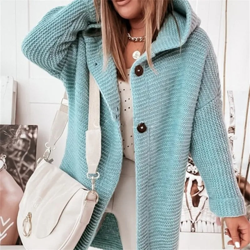 Femme portant un manteau en tricot bleu avec capuche, boutons marron et sac blanc. Mode automne-hiver, style décontracté chic.