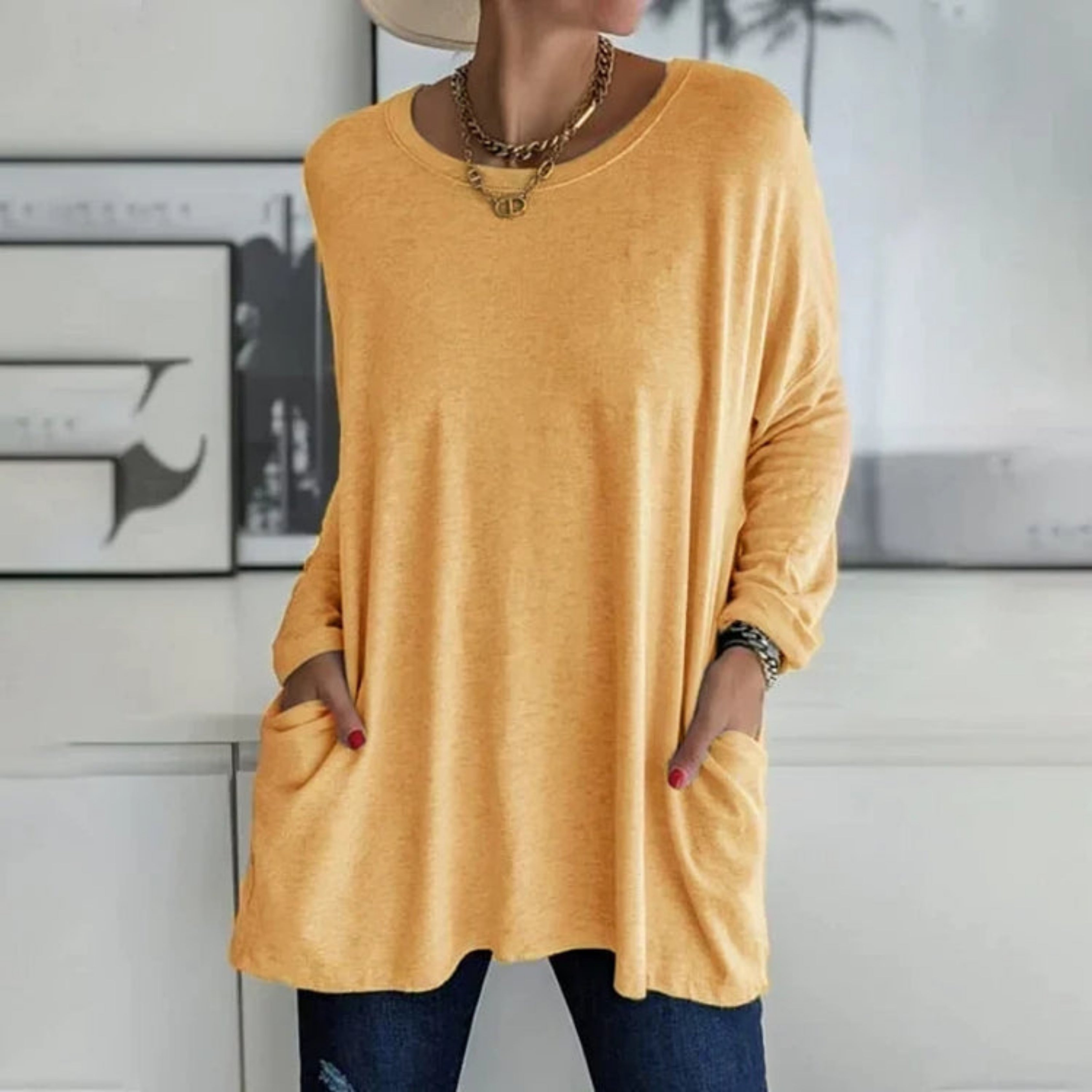 Femme portant un pull ample jaune avec poches, collier doré, chapeau beige, posant devant un mur moderne. Mode décontractée, tendance automne.