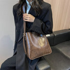 Femme en manteau noir tenant un sac à bandoulière en cuir marron, assise sur un canapé noir. Mode élégante, accessoires tendance, style automne.