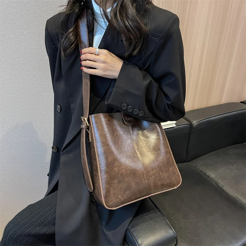 Femme en manteau noir tenant un sac à bandoulière en cuir marron, assise sur un canapé noir. Mode élégante, accessoires tendance, style automne.