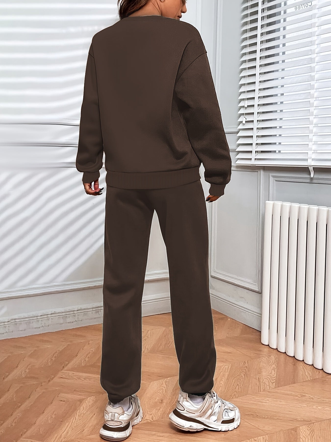 Personne portant un ensemble de survêtement marron, debout dans une pièce lumineuse avec parquet en bois et stores blancs. Mode décontractée, tendance.