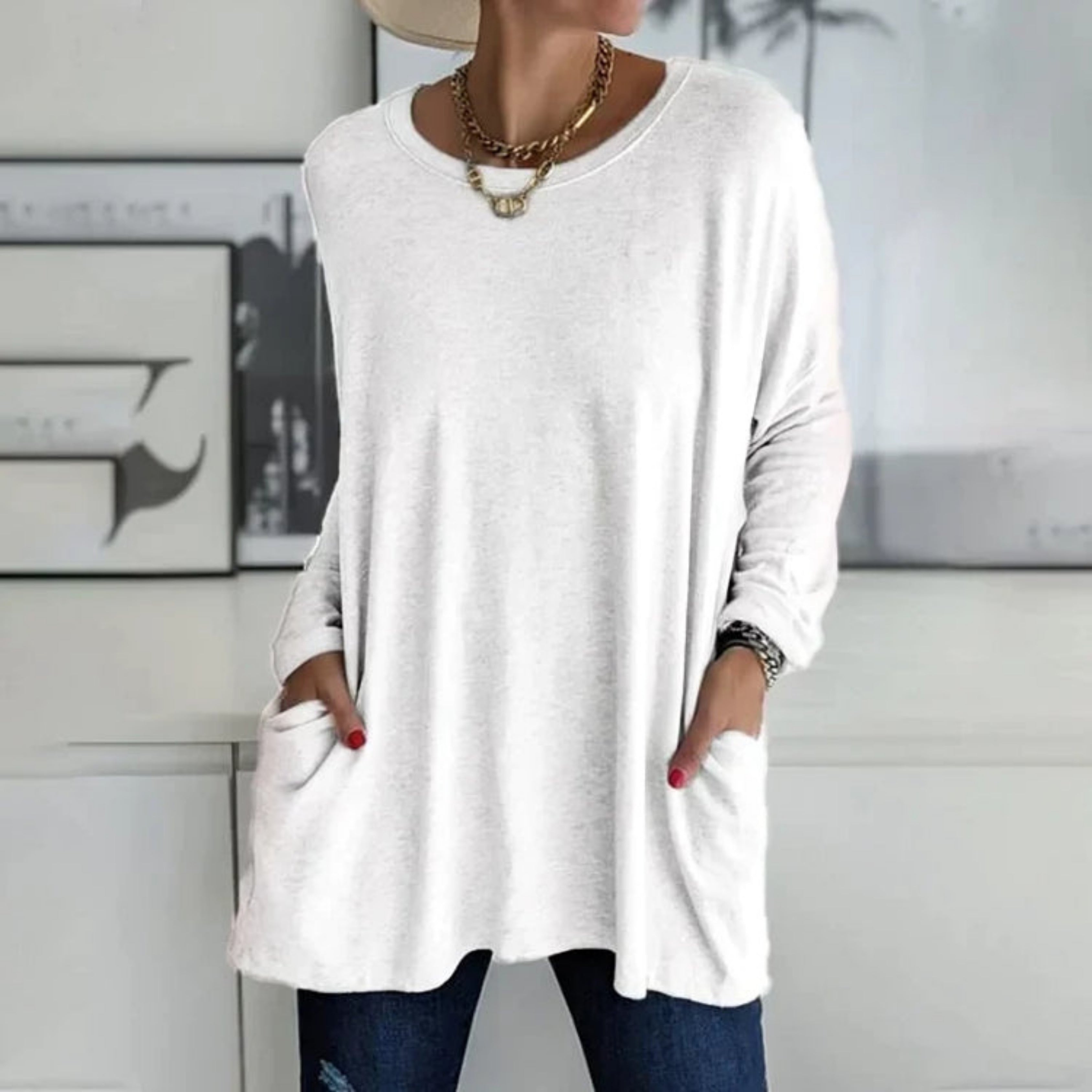 Femme portant un pull blanc ample avec poches, collier doré, et jeans bleu. Mode décontractée, style moderne, tenue automne-hiver.