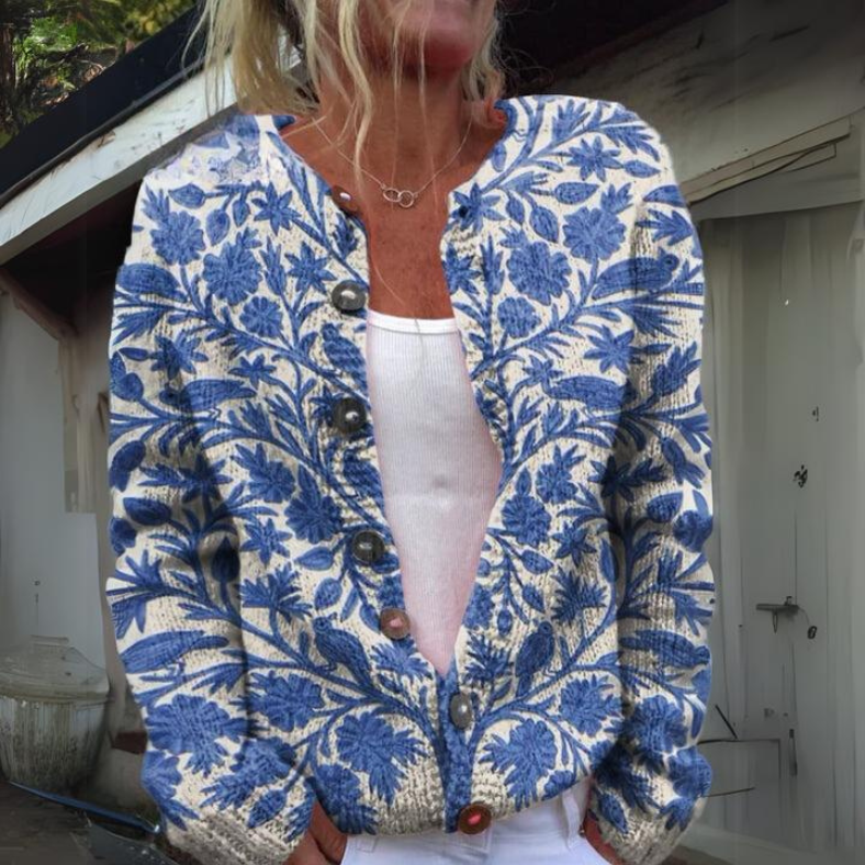 Femme portant un cardigan bleu et blanc à motifs floraux, sur un débardeur blanc, devant une maison. Mode automne, style décontracté, tendance.