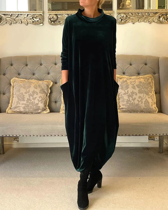Robe longue en velours vert foncé avec poches, portée par une femme debout devant un canapé élégant, décoré de coussins à motifs.