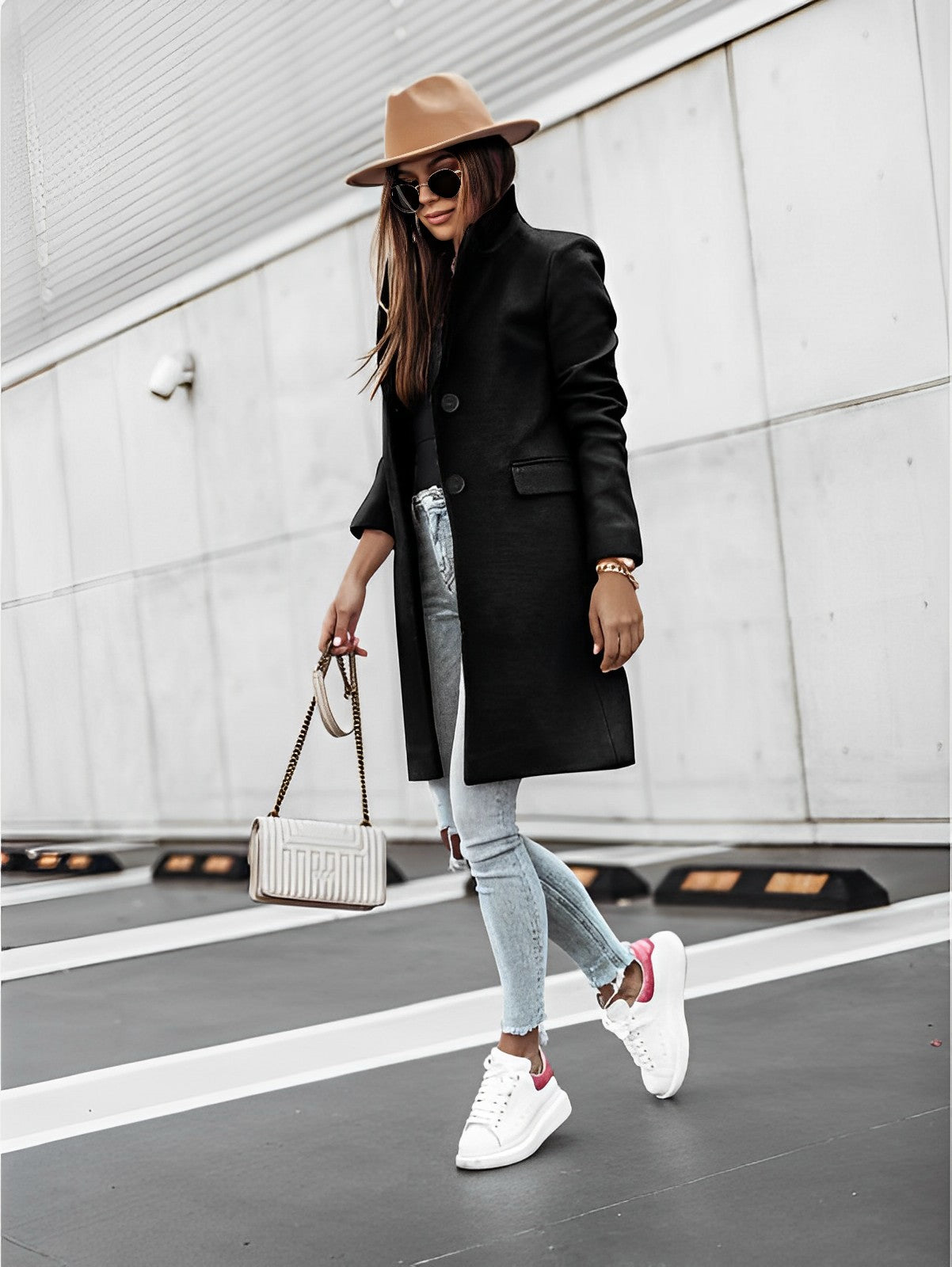 Femme élégante en manteau noir, jeans déchiré, baskets blanches et chapeau beige, tenant un sac à main, posant dans un parking urbain moderne.