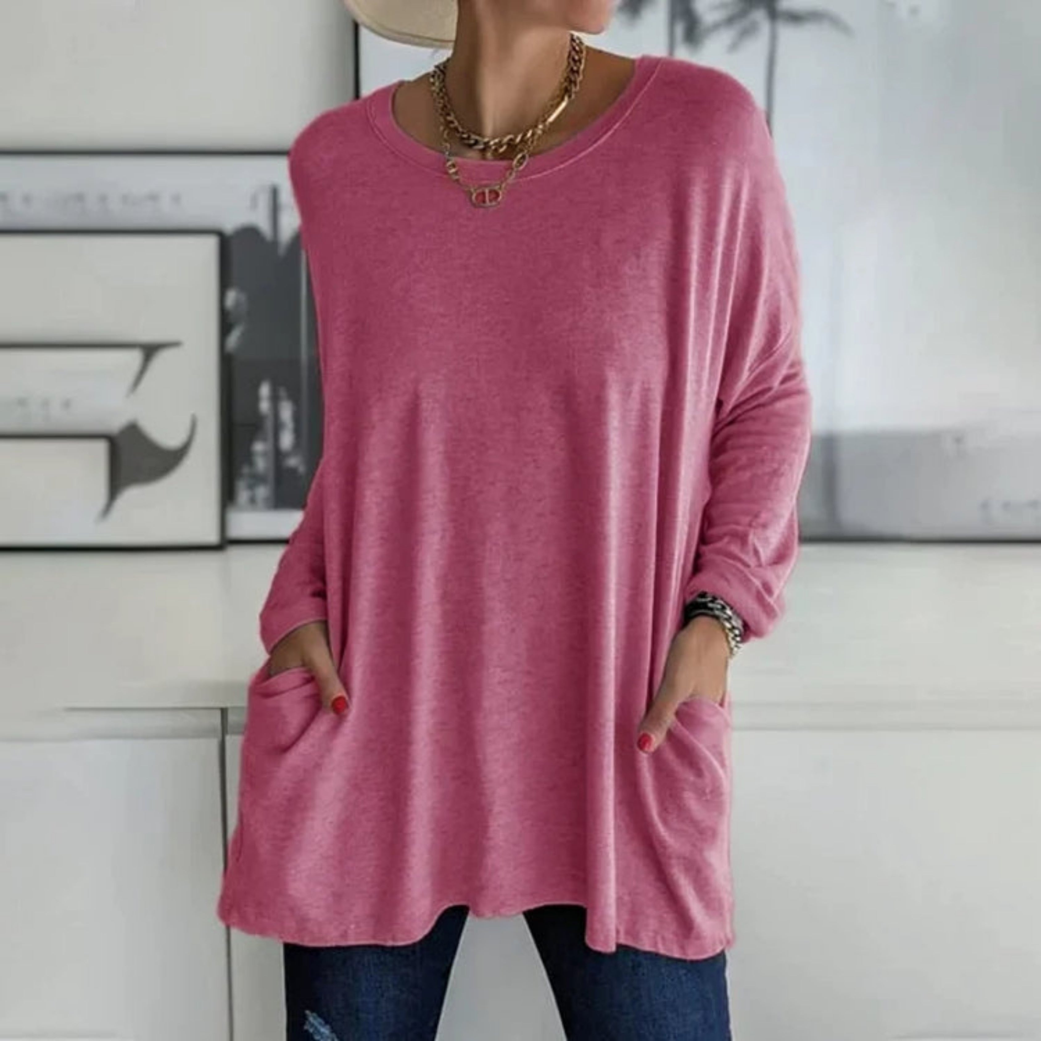 Femme portant un pull rose ample avec poches, collier doré, chapeau beige, posant devant un mur blanc. Mode décontractée, tendance automne.