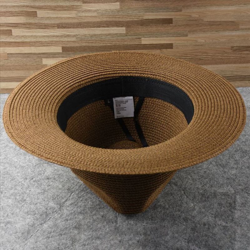 Chapeau de paille marron avec large bord, posé à l'envers sur un sol gris, étiquette visible à l'intérieur. Accessoire mode été, style décontracté.