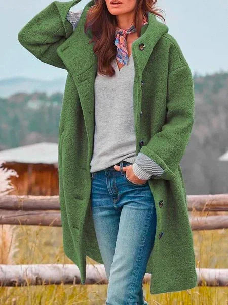 Femme portant un manteau vert en laine, pull gris, jeans, foulard coloré, posant en extérieur. Mode automne-hiver, style décontracté.