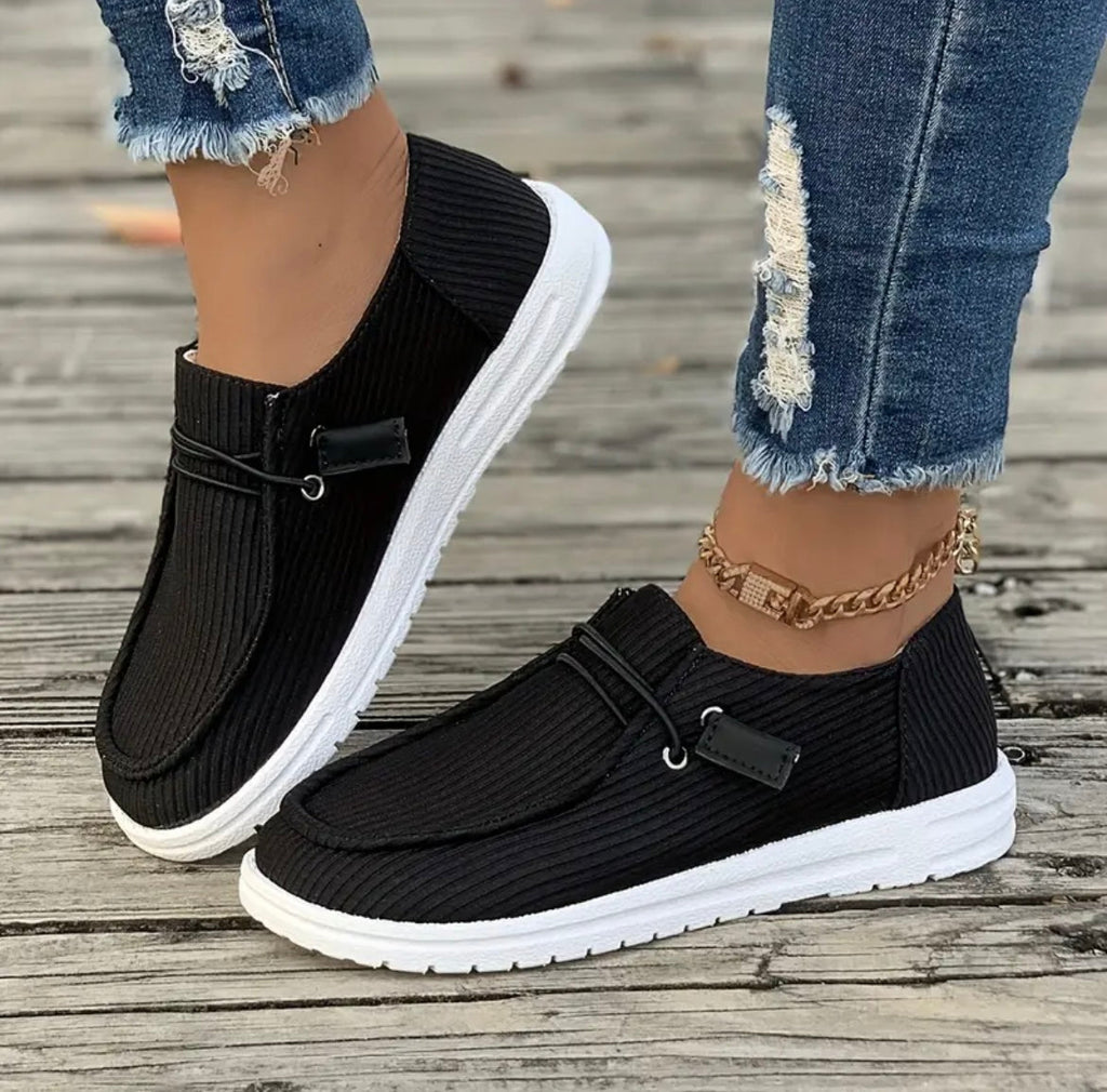 Chaussures décontractées noires pour femmes avec semelles blanches, portées avec un jean déchiré et un bracelet doré, sur fond de bois.