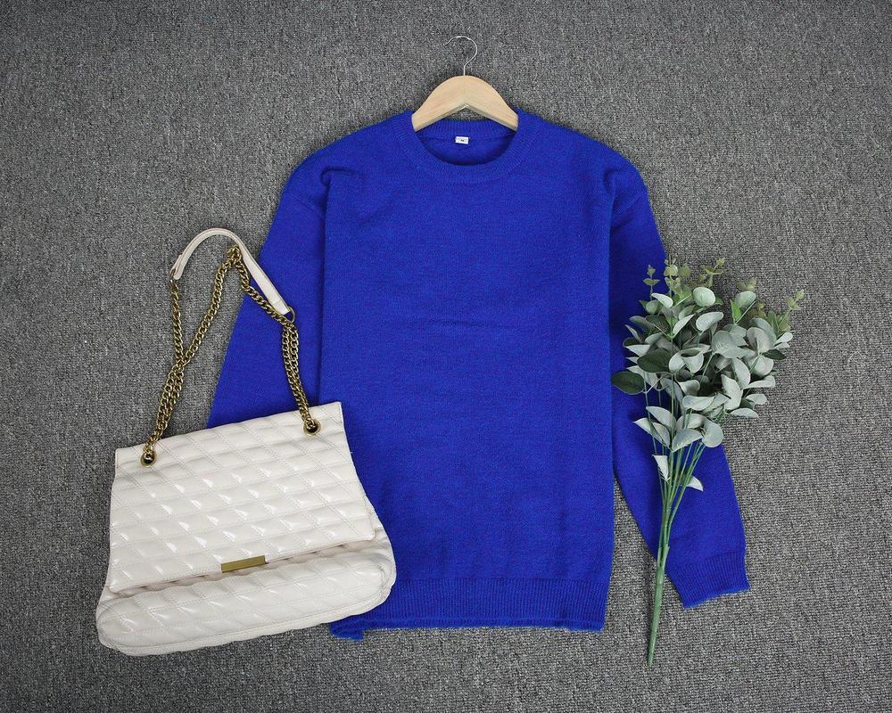 Pull bleu en laine sur cintre, sac à main beige avec chaîne dorée, bouquet de feuilles vertes, posés sur tapis gris. Mode automne-hiver élégante.