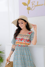 Femme souriante en robe bohème fleurie, chapeau de paille, posant devant un mur blanc. Mode estivale, style bohème chic, accessoires élégants.