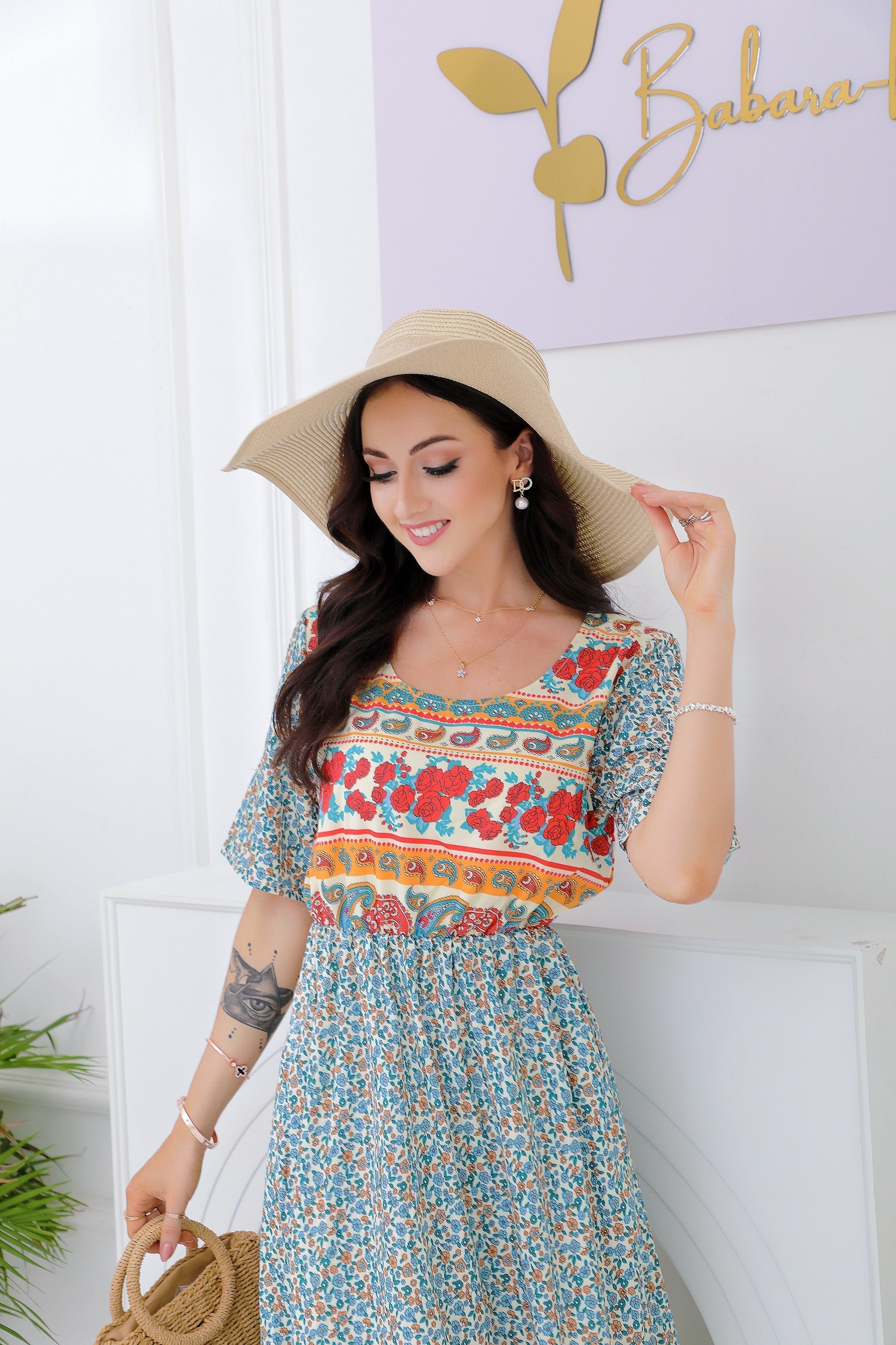 Femme souriante en robe bohème fleurie, chapeau de paille, posant devant un mur blanc. Mode estivale, style bohème chic, accessoires élégants.