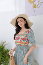 Femme souriante en robe fleurie colorée et chapeau de paille, tenant un sac en osier, posant devant un mur blanc avec plante verte.