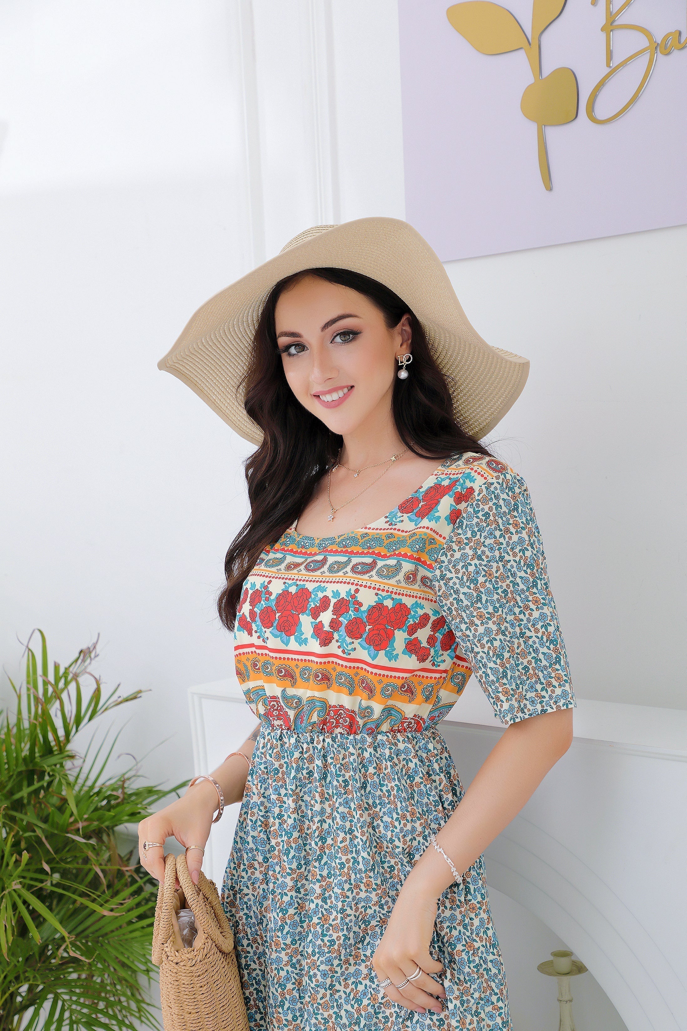 Femme souriante en robe fleurie colorée et chapeau de paille, tenant un sac en osier, posant devant un mur blanc avec plante verte.