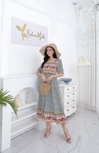 Femme en robe bohème fleurie, chapeau de paille, tenant un sac en osier, posant dans un intérieur élégant avec décoration blanche et plante verte.